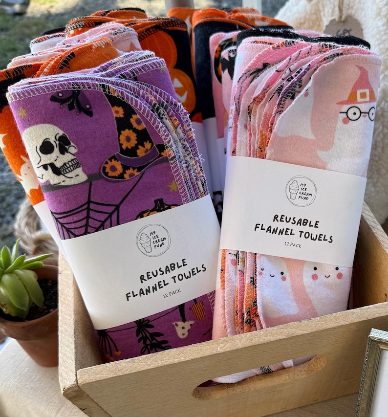 May include: Stacks of reusable flannel towels in a wooden crate. The towels feature Halloween-themed designs, including skulls, witch hats, pumpkins, and ghosts. Each pack is labeled "REUSABLE FLANNEL TOWELS 12 PACK".