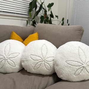 May include: Three white, round decorative pillows with a sand dollar design embroidered in gray thread. The pillows are arranged on a gray sofa with a yellow pillow in the background. The pillows have a soft, textured appearance.