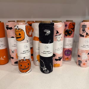 May include: A collection of rolled reusable flannel towels with Halloween-themed patterns. Designs include jack-o'-lanterns, bats, cats in witch hats, ghosts, and skeletons. The towels are in shades of orange, pink, and black.