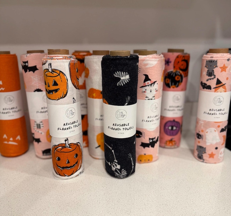 May include: A collection of rolled reusable flannel towels with Halloween-themed patterns. Designs include jack-o'-lanterns, bats, cats in witch hats, ghosts, and skeletons. The towels are in shades of orange, pink, and black.