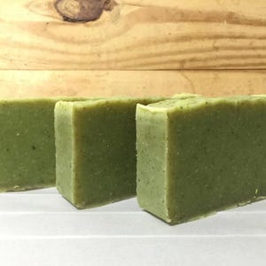 May include: Three green rectangular bars of soap are arranged in a row. The soap has a slightly textured surface and a light green colour. The background is a light-coloured wooden surface.