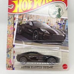 May include: A black Aston Martin One-77 die-cast model car from Hot Wheels. The packaging is designed to look like a postcard, with the words "Hot Wheels" in yellow and red. The card also features the text "United Kingdom" and "Aston Martin One-77".