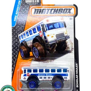 Könnte beinhalten: Ein Matchbox Scholar Hauler Spielzeug-LKW auf seiner Originalverpackung. Der weiß-blaue Schulbus-LKW hat große Geländereifen und die Worte "Scholar Hauler" auf der Seite aufgedruckt. Die Verpackung enthält den Text "MBX Adventure City".