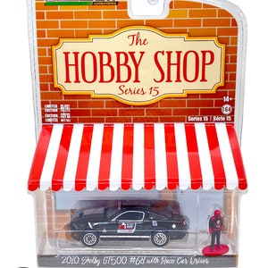 Peut inclure: Une voiture miniature noire 2010 Shelby GT500 #68 avec une figurine de pilote de course dans une vitrine en plastique transparente. La vitrine est décorée d'un auvent rayé rouge et blanc et d'une enseigne qui indique "The Hobby Shop Series 15".
