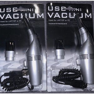 Puede incluir: Dos mini aspiradoras plateadas para portátiles o PC, cada una empaquetada en un blíster transparente. El embalaje incluye el texto "USE MINI VACUUM" y "Used for LAPTOP or PC keyboard/mainboard". Cada aspiradora tiene un cable USB.