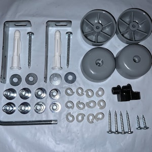 Puede incluir: Un conjunto de componentes de ferretería para muebles, que incluye ruedas grises, soportes metálicos, tornillos, arandelas y un conector de plástico negro. El hardware está dispuesto sobre una superficie blanca, lo que sugiere un kit de reparación o montaje.