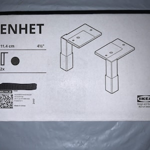 Puede incluir: Caja de cartón blanca con la inscripción "ENHET" y un diagrama de dos patas de mueble metálicas. La caja indica que las patas miden 11,4 cm y 4 1/2 pulgadas. El logotipo de IKEA es visible.