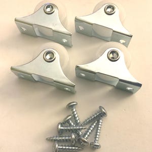 May include: Four silver cabinet rollers with white wheels and mounting plates. A collection of silver screws is also included. These hardware components are designed for furniture repair or construction.