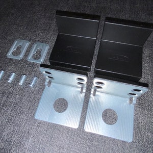 May include: Two black metal brackets with a silver metal bracket attached to each. The brackets have a hole in the centre and are designed for mounting. There are also four small silver screws and two silver metal pieces with a hole in the centre.