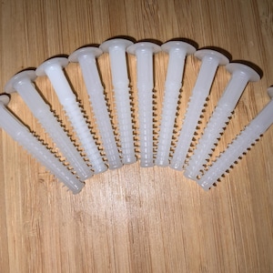 May include: A collection of ten white plastic wall anchors, arranged in a fan shape. Each anchor has a round head and a ridged shaft, designed for secure installation in drywall or similar materials. The anchors are on a wooden surface.