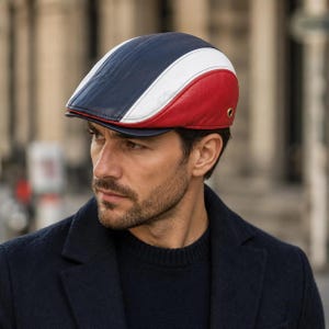May include: A flat cap constructed from panels of navy blue, white, and red leather. The cap features a structured brim and a small metallic grommet detail on the side panel, showcasing a multi-colored design with a smooth, polished finish.