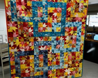 May include: A colorful quilt with a patchwork design featuring red, yellow, blue, and orange squares and triangles. The quilt is hanging on a wall.