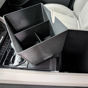 May include: A black, textured car console organizer with multiple compartments. The organizer is designed to fit inside a car's center console, providing storage and organization. The organizer is made of a durable material and is designed to fit a variety of car models.