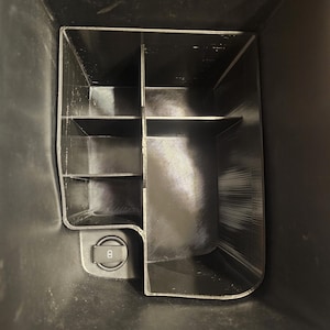 May include: A black plastic organizer with multiple compartments for storing small items. The organizer is designed to fit in a car's center console and has a built-in power outlet.