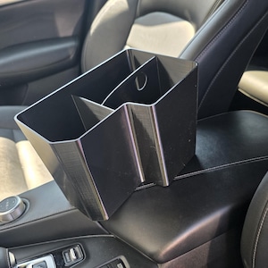 Op de afbeelding: Een zwarte auto-console-organizer met meerdere compartimenten. De organizer is ontworpen om in de middenconsole van een voertuig te passen. De afbeelding toont de organizer in een auto-interieur, met de stoelen en het dashboard zichtbaar.