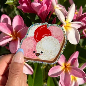 Peut inclure: Un autocollant de pain grill&eacute; de dessin anim&eacute; rose et blanc avec un nappage de confiture de fraises rouge et un visage souriant. L'autocollant a une bordure scintillante et est tenu dans une main.
