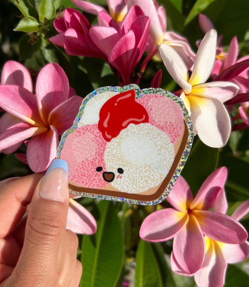 Peut inclure: Un autocollant de pain grill&eacute; de dessin anim&eacute; rose et blanc avec un nappage de confiture de fraises rouge et un visage souriant. L'autocollant a une bordure scintillante et est tenu dans une main.