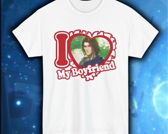 May include: White t-shirt with a graphic design. The design features a red heart-shaped frame with the words "I My Boyfriend" and an illustration of a person inside the heart. The t-shirt is made of a soft material.