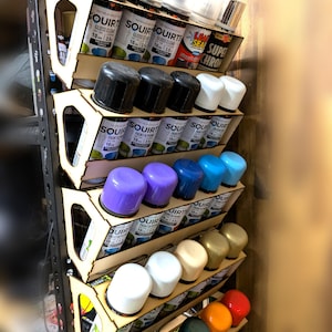May include: A wooden spray paint organizer filled with various spray paint cans. The cans are arranged in angled slots, showcasing a variety of colors, including black, white, purple, blue, and green. The brand name "SQUIRT" is visible on the cans.
