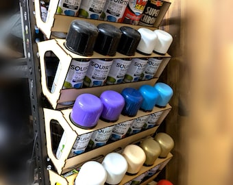 May include: A wooden spray paint organizer filled with various spray paint cans. The cans are arranged in angled slots, showcasing a variety of colors, including black, white, purple, blue, and green. The brand name "SQUIRT" is visible on the cans.