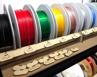 May include: A display of colorful 3D printer filament spools. The spools are arranged in a row, showcasing various colors including black, red, green, yellow, and blue. Each spool is labeled with a tag indicating the filament type, such as "PLA" and "PETG".
