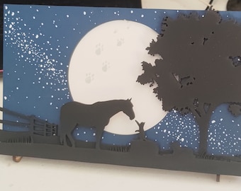 May include: A black silhouette of a horse and a cat standing in a field under a full moon with a black silhouette of a tree in the background. The sky is blue with white stars.