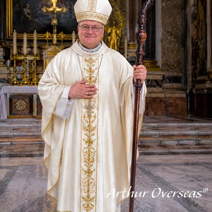 May include: A bishop wearing a cream-colored liturgical chasuble with gold embroidery, a matching miter, and a pectoral cross. The bishop holds a tall, dark brown wooden crozier with an ornate, carved spiral top. The text Arthur Overseas is visible.