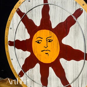 Pode incluir: Um escudo redondo de madeira com um desenho de sol. O sol &eacute; laranja com um rosto, rodeado por raios vermelhos, sobre um fundo branco e com gr&atilde;o de madeira. Um anel de metal est&aacute; preso na frente. O escudo tem uma borda de couro.