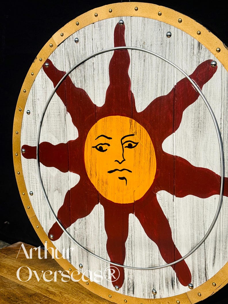 Pode incluir: Um escudo redondo de madeira com um desenho de sol. O sol &eacute; laranja com um rosto, rodeado por raios vermelhos, sobre um fundo branco e com gr&atilde;o de madeira. Um anel de metal est&aacute; preso na frente. O escudo tem uma borda de couro.