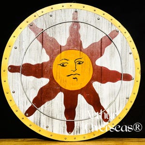 Pode incluir: Um escudo de madeira redondo com um desenho de sol. O sol tem um rosto amarelo e raios vermelhos sobre um fundo branco e castanho. O escudo tem uma borda dourada e um anel de metal prateado.