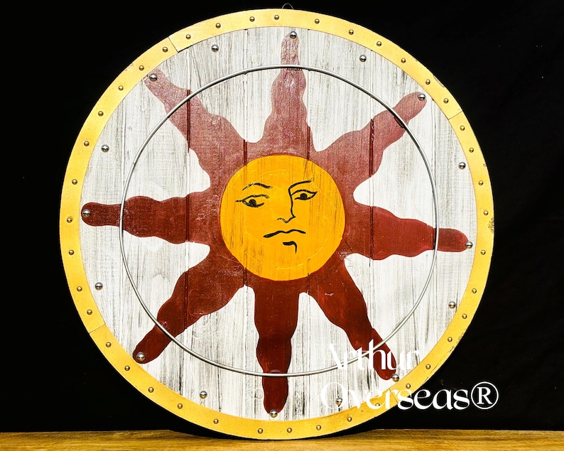 Pode incluir: Um escudo de madeira redondo com um desenho de sol. O sol tem um rosto amarelo e raios vermelhos sobre um fundo branco e castanho. O escudo tem uma borda dourada e um anel de metal prateado.