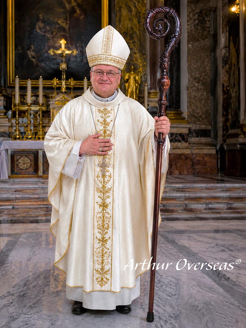 May include: A bishop wearing a cream-colored liturgical chasuble with gold embroidery, a matching miter, and a pectoral cross. The bishop holds a tall, dark brown wooden crozier with an ornate, carved spiral top. The text Arthur Overseas is visible.