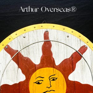Pode incluir: Escudo de madeira com um desenho de sol. O escudo apresenta uma borda amarela, raios de sol brancos e vermelhos e uma face de sol amarela. O texto "Arthur Overseas" est&aacute; no topo. Um anel de metal est&aacute; preso ao escudo.