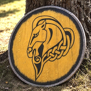 Op de afbeelding: Een rond houten schild met een geel geschilderd oppervlak en een zwart geschilderd paardenhoofdontwerp. De paardenillustratie bevat ingewikkelde knooppatronen. Het schild heeft een donkere, verweerde metalen rand aan de buitenkant.