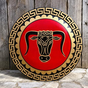 Könnte beinhalten: Eine kreisförmige Wandplatte mit einem zentralen roten Feld und einer schwarzen Stierkopf-Illustration. Das Design ist von einem gewellten goldenen Rand und einem äußeren schwarzen Ring mit einem goldenen griechischen Mäandermuster umgeben.