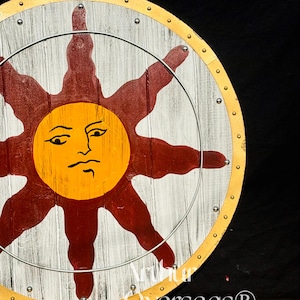 Pode incluir: Um escudo redondo de madeira com um desenho de sol. O sol &eacute; amarelo com um rosto, rodeado por raios vermelhos. O escudo tem um fundo branco e cinza, um anel de metal e uma borda dourada. O texto "Arthur Overseas&reg;" &eacute; vis&iacute;vel.
