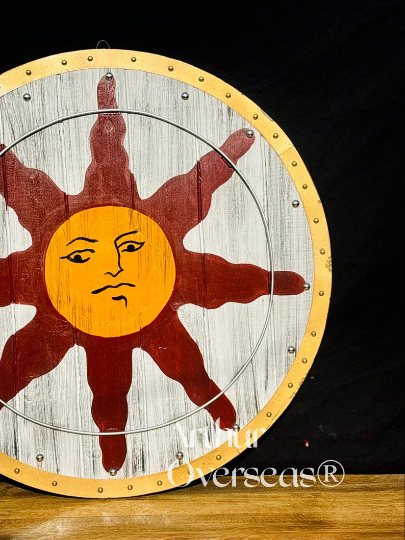 Pode incluir: Um escudo redondo de madeira com um desenho de sol. O sol &eacute; amarelo com um rosto, rodeado por raios vermelhos. O escudo tem um fundo branco e cinza, um anel de metal e uma borda dourada. O texto "Arthur Overseas&reg;" &eacute; vis&iacute;vel.