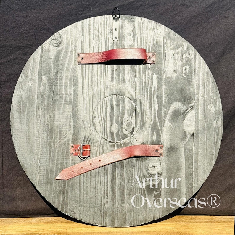 Pode incluir: Um escudo de madeira redondo com um acabamento cinza lavado. Possui correias de couro vermelho e uma argola de metal para pendurar. O escudo tem um aspecto envelhecido, com gr&atilde;o de madeira vis&iacute;vel e o texto "Arthur Overseas&reg;".