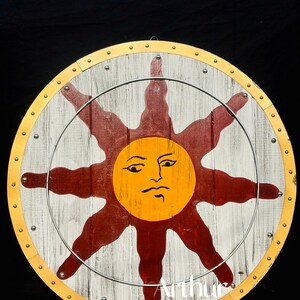 Pode incluir: Um escudo redondo de madeira com fundo branco e cinza, apresentando um grande desenho de sol em laranja e vermelho. O escudo tem uma borda dourada e &eacute; fixado com rebites de metal. O texto "Arthur Overseas&reg;" &eacute; vis&iacute;vel.