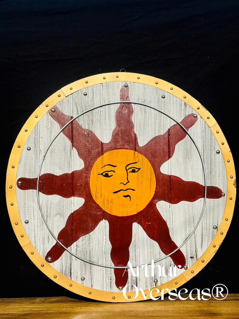 Pode incluir: Um escudo redondo de madeira com fundo branco e cinza, apresentando um grande desenho de sol em laranja e vermelho. O escudo tem uma borda dourada e &eacute; fixado com rebites de metal. O texto "Arthur Overseas&reg;" &eacute; vis&iacute;vel.
