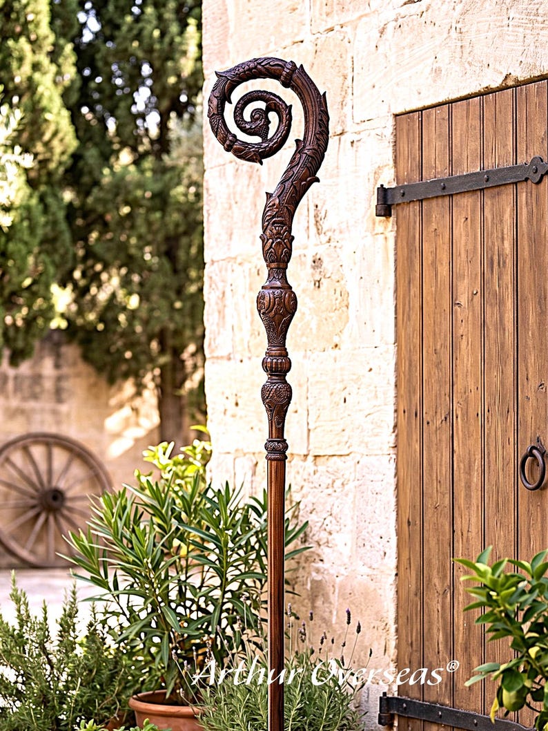 May include: A tall, dark brown wooden crozier featuring an intricately carved spiral top with leaf-like patterns and a textured, segmented shaft. The staff displays detailed craftsmanship with ornate carvings along the neck and handle sections.