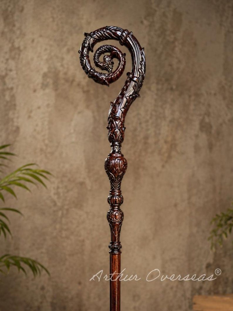 May include: A dark brown wooden staff featuring an intricate, carved spiral top with dragon-like motifs and detailed scrollwork. The shaft includes decorative, rounded nodes below the head, with the text Arthur Overseas printed at the bottom.