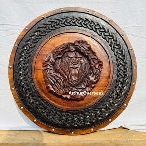 Puede incluir: Un escudo redondo de madera con una cara de oso tallada en el centro. El escudo está decorado con un diseño de nudo celta alrededor del borde y tiene tachuelas de metal alrededor del perímetro.