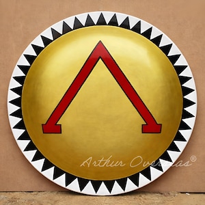 Könnte beinhalten: Ein runder Schild mit goldenem Zentrum und einem roten Lambda-Symbol. Der Schild hat einen weißen Rand mit schwarzen Dreiecken. Der Text "Arthur Overseas" ist unten rechts in Kursivschrift geschrieben.