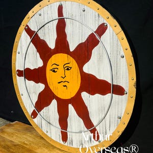Pode incluir: Um escudo redondo de madeira com fundo branco e um grande desenho de sol estilizado em vermelho e amarelo. O escudo tem um anel de metal e &eacute; adornado com rebites dourados e prateados. O texto "Arthur Overseas&reg;" &eacute; vis&iacute;vel.