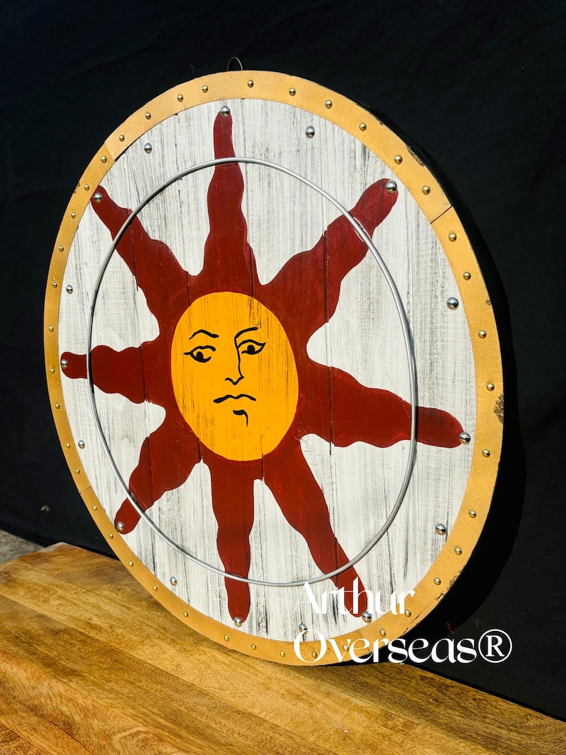 Pode incluir: Um escudo redondo de madeira com fundo branco e um grande desenho de sol estilizado em vermelho e amarelo. O escudo tem um anel de metal e &eacute; adornado com rebites dourados e prateados. O texto "Arthur Overseas&reg;" &eacute; vis&iacute;vel.