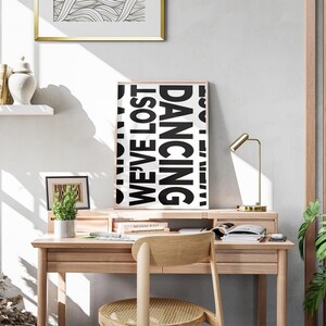 May include: A black and white poster with the text 'Dancing We've Lost' hanging on a wall above a wooden desk. The desk has a gold lamp, a potted plant, and a chair with a woven seat. There are books and a white object on the desk.