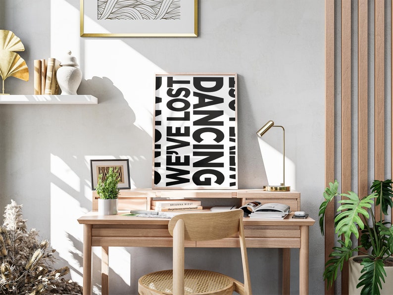 May include: A black and white poster with the text 'Dancing We've Lost' hanging on a wall above a wooden desk. The desk has a gold lamp, a potted plant, and a chair with a woven seat. There are books and a white object on the desk.