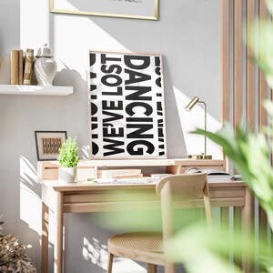 May include: A black and white poster with the words 'WE'VE LOST' and 'DANCING' in large, bold letters. The poster is hanging on a white wall above a wooden desk with a chair, a lamp, and a potted plant. The desk is in a room with a wooden wall and a window with natural light coming through.