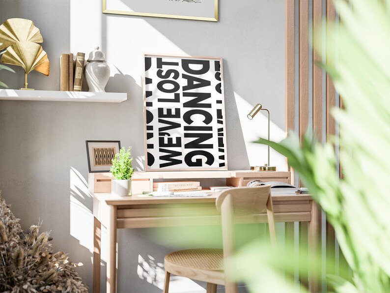 May include: A black and white poster with the words 'WE'VE LOST' and 'DANCING' in large, bold letters. The poster is hanging on a white wall above a wooden desk with a chair, a lamp, and a potted plant. The desk is in a room with a wooden wall and a window with natural light coming through.